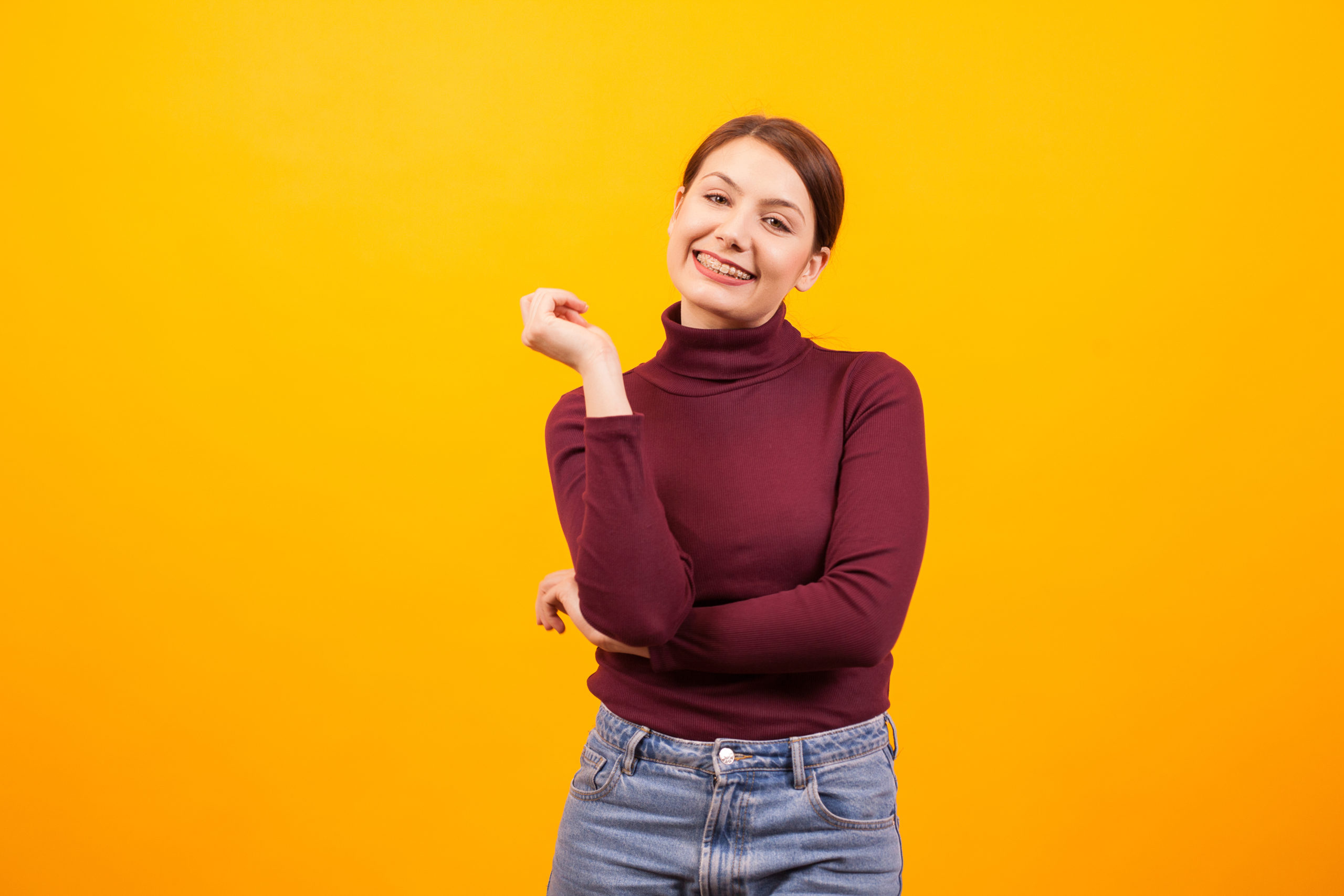 Young woman wearing a red turtleneck and jeans smiling, showing off her braces and standing in front of a yellow background